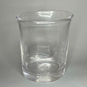 May include: A clear, cylindrical drinking glass with a slightly flared rim. The glass is made of transparent material, allowing light to pass through. The base is a solid, flat circle. The glass is empty and sits on a gray surface.