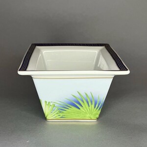 May include: Square white ceramic bowl with a blue and green palm frond design. The rim features a dark blue Greek key pattern. A gold trim accents the top and base of the bowl. The bowl has a light blue background.