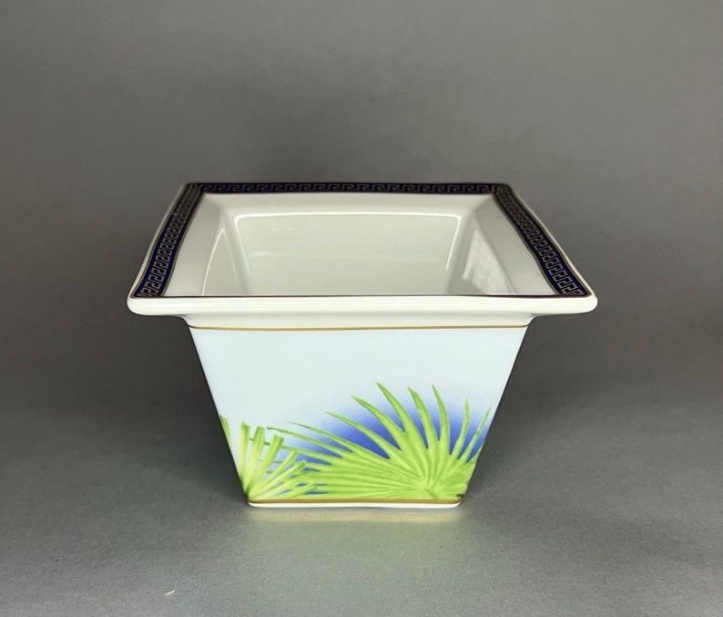 May include: Square white ceramic bowl with a blue and green palm frond design. The rim features a dark blue Greek key pattern. A gold trim accents the top and base of the bowl. The bowl has a light blue background.