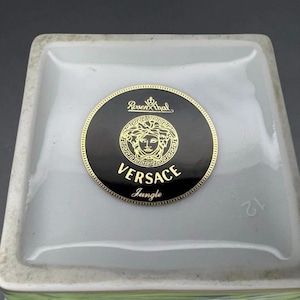 May include: Close-up of a square, white ceramic object with a black and gold Versace Jungle logo. The logo features a Medusa head and the Rosenthal brand name. The number 12 is visible on the side.