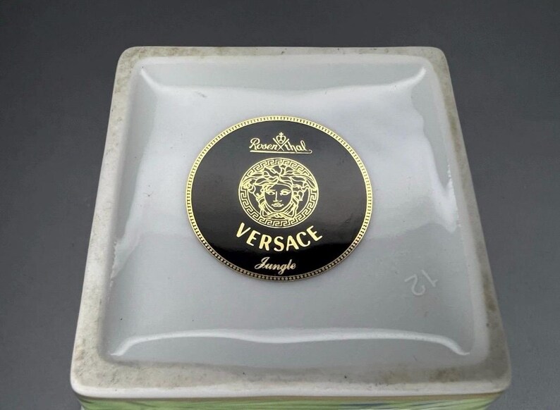 May include: Close-up of a square, white ceramic object with a black and gold Versace Jungle logo. The logo features a Medusa head and the Rosenthal brand name. The number 12 is visible on the side.
