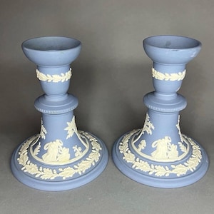 May include: Two blue ceramic candlesticks with white relief designs. Each candlestick features a circular base, a central column, and a top cup. The bases are decorated with a white relief of figures and foliage. The candlesticks are approximately 6 inches tall.