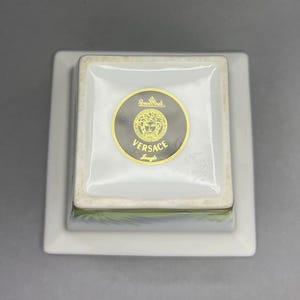 May include: A square, white porcelain dish with a smaller square dish on top. The top dish has a gold and black Versace logo. The logo features the Versace name and a Medusa head. The dish is a luxury item.