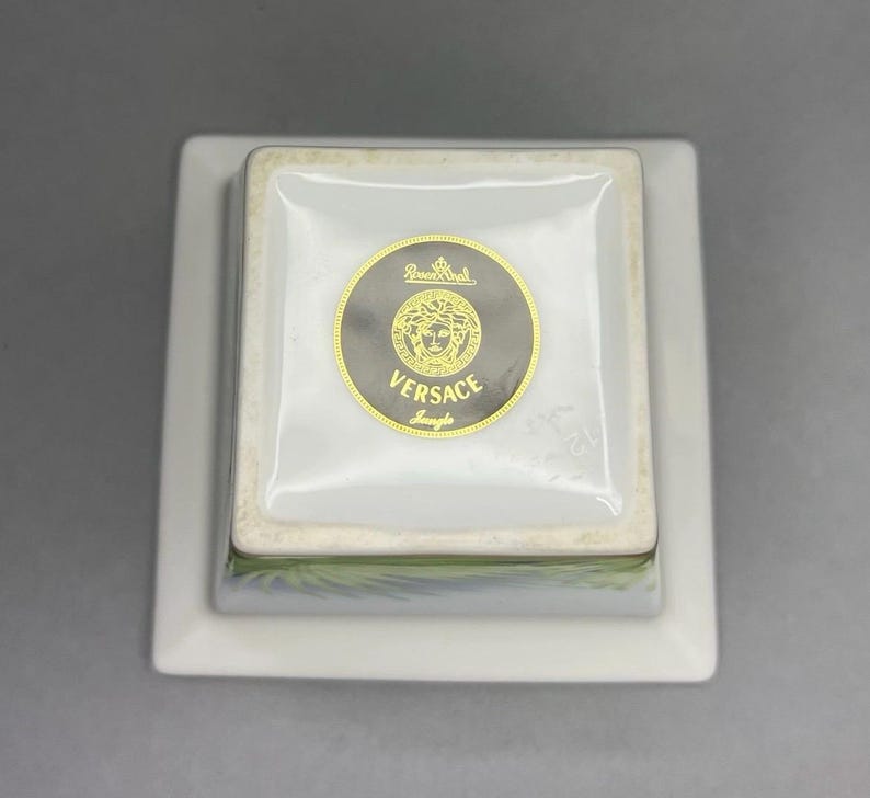 May include: A square, white porcelain dish with a smaller square dish on top. The top dish has a gold and black Versace logo. The logo features the Versace name and a Medusa head. The dish is a luxury item.