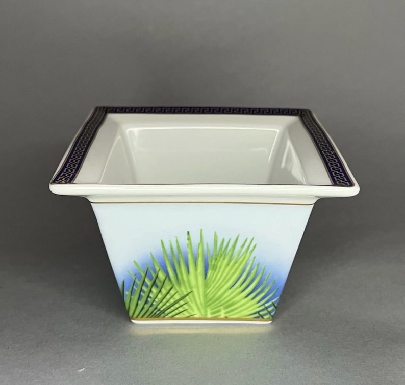 May include: A square, white ceramic bowl with a blue and green palm frond design. The rim is decorated with a dark blue Greek key pattern. A gold trim accents the top and bottom edges of the bowl.