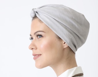 Op de afbeelding: Een lichtgrijze turban-stijl hoofdband gemaakt van stof. De band is bovenop het hoofd in een knoop geknoopt.