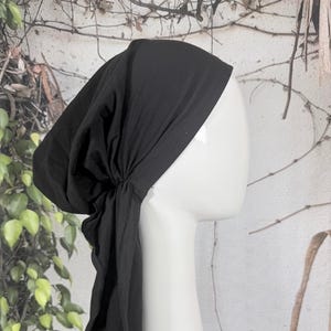 May include: A black head covering, likely a bonnet or scarf, is draped over a white mannequin head. The fabric is gathered at the back and hangs down. The background features greenery and a neutral-toned wall.