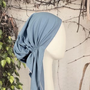 Może przedstawiać: Jasnoniebieska chusta na głowę, prawdopodobnie hidżab lub szal, jest prezentowana na białej głowie manekina. Materiał jest marszczony z tyłu i opada na szyję. Krawędzie wykończone białymi szwami. Tło przedstawia zieleń i neutralne odcienie.