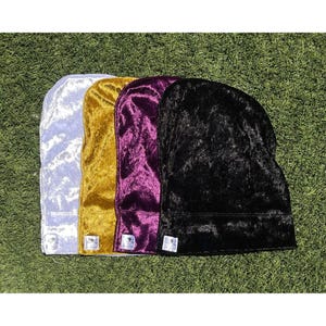 May include: Four velvet durags in a row, each in a different color: light blue, gold, purple, and black. The durags are arranged on a green surface. Each durag has a small white tag at the bottom.