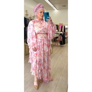 Pode incluir: Um vestido maxi de estampa floral rosa e branco com gola alta e mangas compridas. O vestido tem uma saia em camadas e um corpete ajustado. A modelo está usando um lenço rosa na cabeça.