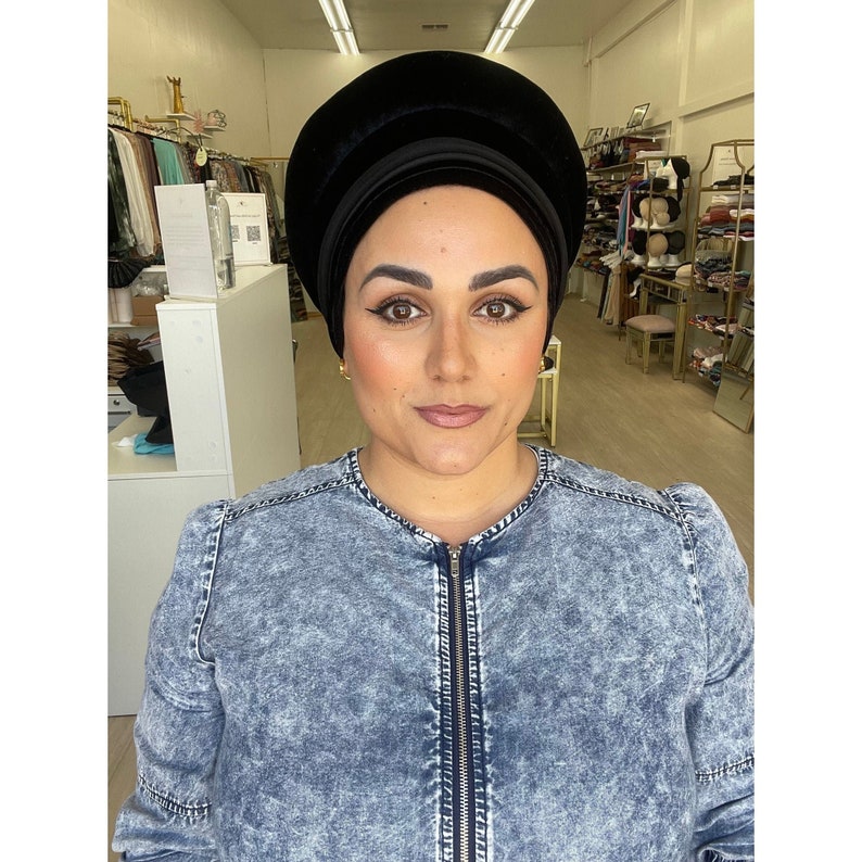 May include: A woman wearing a black velvet turban and a denim jacket with a zipper. The turban is a rounded, layered design. The jacket is a light blue wash with a zip-up front.
