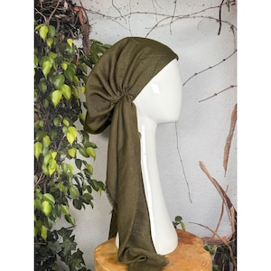 May include: Olive green head covering with long, flowing fabric. The head wrap is draped over a white mannequin head, showcasing its design. The fabric has a textured appearance, and the style is reminiscent of a turban.