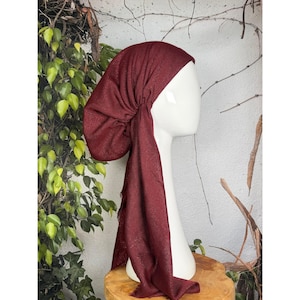 May include: A burgundy head covering with a draped design, displayed on a white mannequin head. The fabric has a subtle shimmer and a fringed edge. The head covering is likely a turban or head wrap.
