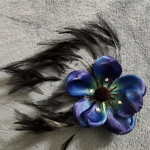 Könnte beinhalten: Ein blau-violettes Haaraccessoire in Form einer Blume mit schwarzen Federn. Die Blume hat fünf Blütenblätter mit Farbverlauf und kleinen, funkelnden Verzierungen. Die Federn sind auf der Rückseite der Blume befestigt.