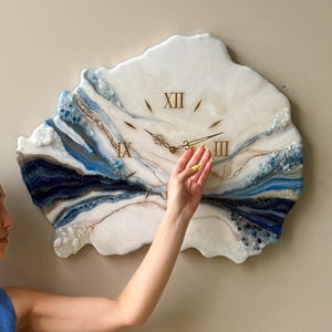 Puede incluir: Un reloj de pared decorativo con un diseño de resina blanca, azul y dorada. La esfera del reloj tiene números romanos y agujas doradas. El reloj está montado en una pared beige. Una persona está de pie cerca del reloj.