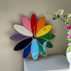 Może przedstawiać: Kolorowy zegar ścienny w kształcie stokrotki ze złotą ramą. Tarcza zegara ma płatki w różnych kolorach, w tym różowym, czerwonym, pomarańczowym, żółtym, zielonym, niebieskim i fioletowym. Wskazówki zegara są złote.