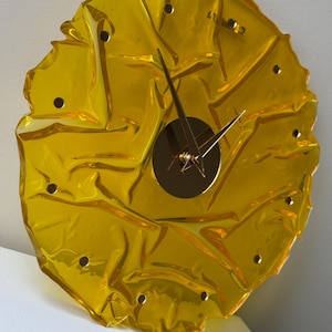 May include: A distinctive, yellow wall clock with a textured, crumpled design. The clock has a gold-coloured centre and hands, and small, evenly spaced holes around the edge. The clock's design is modern and visually striking.