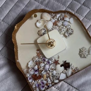 Könnte beinhalten: Eine dekorative Uhr mit cremefarbenem Zifferblatt, verziert mit Muscheln, Perlen und einem kleinen Boot. Die Uhr hat goldfarbene Zeiger und einen goldfarbenen Rand. Das Zifferblatt ist quadratisch und die Zeiger zeigen auf 10:08.