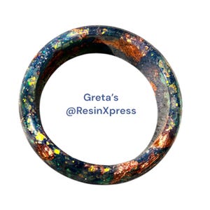 May include: A resin bangle bracelet with a dark blue base, accented with gold and green flecks. The bracelet has a circular shape and features the text "Greta's @ResinXpress".