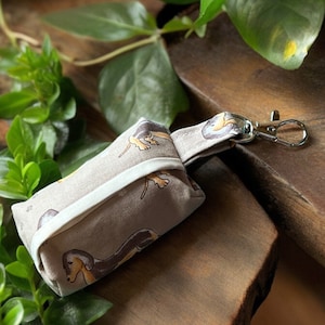 Może przedstawiać: Beżowy dozownik na worki na psie odchody z powtarzającym się wzorem jamnika. Dozownik ma białe wykończenie i srebrny metalowy klips do mocowania do smyczy. Zielone liście otaczają dozownik.