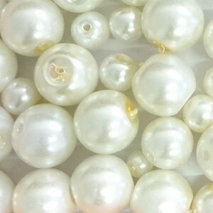 May include: A collection of round, off-white faux pearls of varying sizes. Each bead features a central hole for stringing, with a smooth, lustrous finish that reflects light. The pearls are arranged in a dense cluster, showcasing their spherical shape.