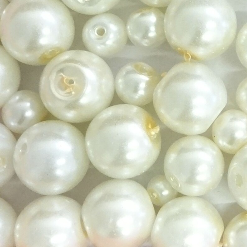 May include: A collection of round, off-white faux pearls of varying sizes. Each bead features a central hole for stringing, with a smooth, lustrous finish that reflects light. The pearls are arranged in a dense cluster, showcasing their spherical shape.