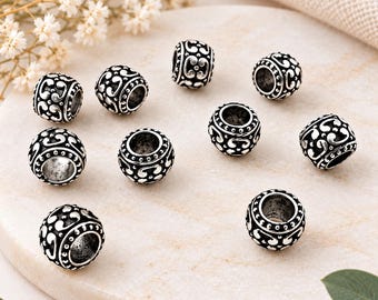 May include: A collection of nine round metal beads featuring an oxidised silver finish with intricate embossed scrollwork patterns and a central hole for threading. Each bead displays a consistent dark recessed design and raised metallic details.