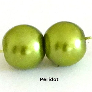 May include: Two round, lime green beads with a smooth, lustrous finish, each featuring a central hole for stringing. The word Peridot is printed in black text below the beads, identifying the color of these spherical craft components.
