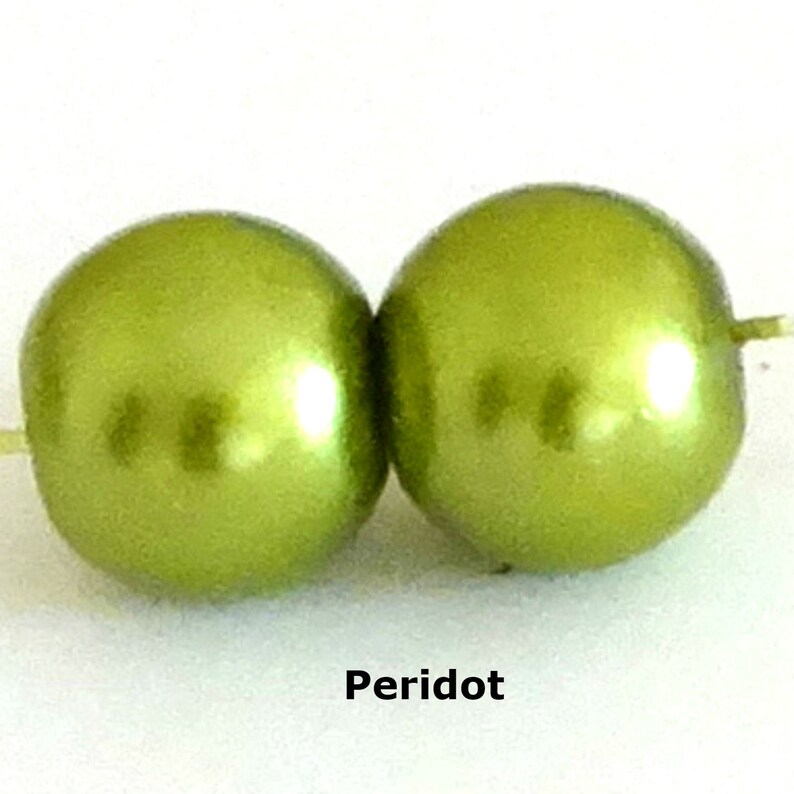 May include: Two round, lime green beads with a smooth, lustrous finish, each featuring a central hole for stringing. The word Peridot is printed in black text below the beads, identifying the color of these spherical craft components.