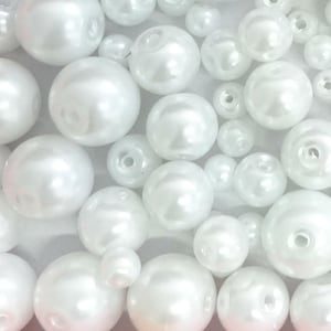 May include: A collection of white, spherical faux pearl beads of varying sizes. Each bead features a smooth, glossy finish and a central hole for threading. The beads are arranged in a dense cluster, showcasing their reflective surfaces and round shapes.