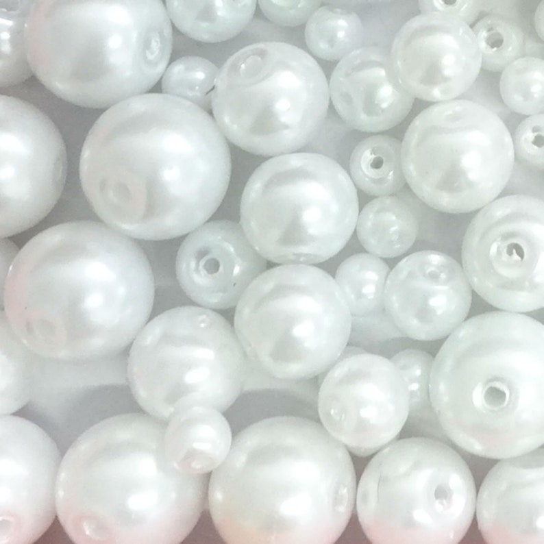 May include: A collection of white, spherical faux pearl beads of varying sizes. Each bead features a smooth, glossy finish and a central hole for threading. The beads are arranged in a dense cluster, showcasing their reflective surfaces and round shapes.