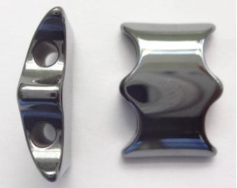 Op de afbeelding: Twee metalen, donkergrijze, gepolijste kralen met een glad, reflecterend oppervlak. Eén kraal is van opzij te zien en onthult twee ronde gaten, terwijl de andere van voren te zien is, met een gebogen, zandlopervormige vorm met uitlopende randen.