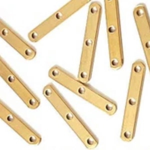 Puede incluir: Un grupo de 12 tiras de metal de color dorado con tres agujeros cada una. Las tiras están dispuestas en un patrón aleatorio sobre una superficie blanca.