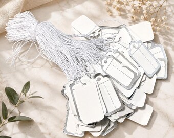 May include: A bundle of small, rectangular white paper tags with silver metallic edges. Each tag features a pre-punched hole at the top and is attached to a long, thin, white string. The tags are grouped together by the strings in a cluster.