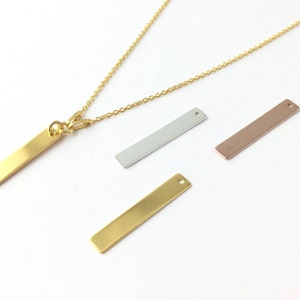 Puede incluir: Un collar de cadena de oro con un colgante de barra de oro largo y delgado. Se muestran tres colgantes de barra adicionales en la imagen: uno plateado, uno dorado y uno de oro rosa.