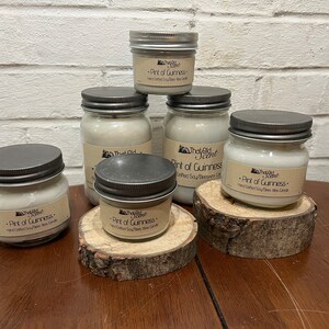 May include: Five glass jars with silver lids containing soy wax candles. The jars are arranged on wooden slices. The labels on the jars read "That Old Scent" and "Pint of Guinness - Hand Crafted Soy/Beeswax Wax Candle".