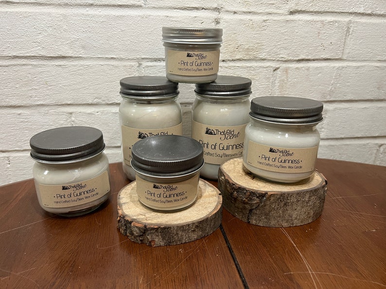 May include: Five glass jars with silver lids containing soy wax candles. The jars are arranged on wooden slices. The labels on the jars read "That Old Scent" and "Pint of Guinness - Hand Crafted Soy/Beeswax Wax Candle".