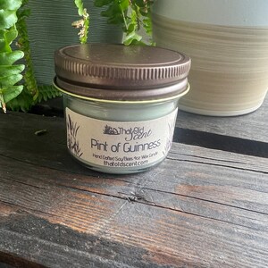 May include: A small glass jar candle with a brown lid. The label reads "That Old Scent" and "Pint of Guinness" with the text "Hand Crafted Soy/Bees 4oz Wax Candle thatoldscent.com".