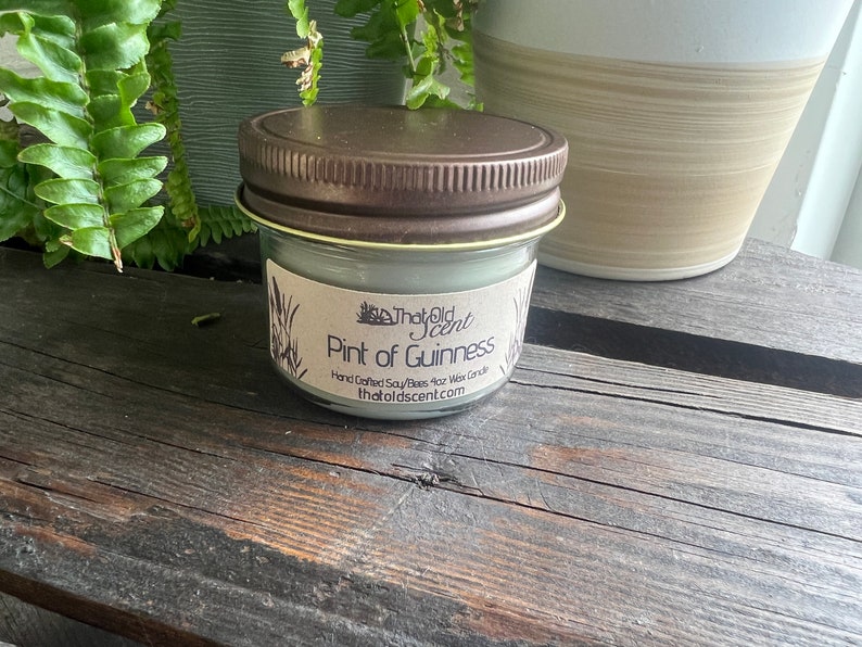 May include: A small glass jar candle with a brown lid. The label reads "That Old Scent" and "Pint of Guinness" with the text "Hand Crafted Soy/Bees 4oz Wax Candle thatoldscent.com".