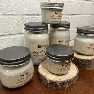 May include: Five glass jars with silver lids containing soy wax candles. The jars are labeled "The Old Jar" and "Pint of Guinness" with the text "Hand Crafted Soy Wax" on each jar. The jars are sitting on wooden rounds.