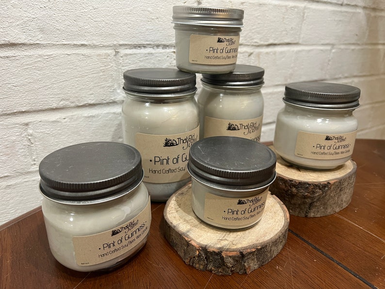 May include: Five glass jars with silver lids containing soy wax candles. The jars are labeled "The Old Jar" and "Pint of Guinness" with the text "Hand Crafted Soy Wax" on each jar. The jars are sitting on wooden rounds.