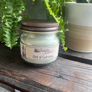 May include: A glass jar candle with a brown lid and a label that reads "That Old Scent Pint of Guinness". The candle is a light green color and is sitting on a wooden surface.