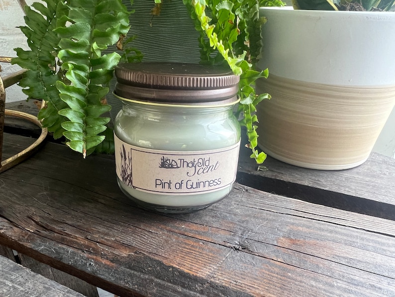 May include: A glass jar candle with a brown lid and a label that reads "That Old Scent Pint of Guinness". The candle is a light green color and is sitting on a wooden surface.