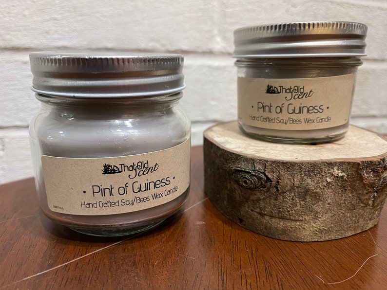 May include: Two glass jars with silver lids containing soy and beeswax candles. The labels on the jars read "That Old Scent" and "Pint of Guiness. Hand Crafted Soy/Bees Wax Candle."