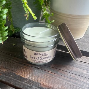 May include: A green soy wax candle in a clear glass jar with a brown lid. The candle is labeled "The Old Scent" and "Pint of Guinness". Hand-crafted soy-based wax candle. TheOldScent.com