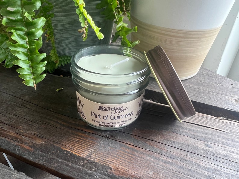 May include: A green soy wax candle in a clear glass jar with a brown lid. The candle is labeled "The Old Scent" and "Pint of Guinness". Hand-crafted soy-based wax candle. TheOldScent.com