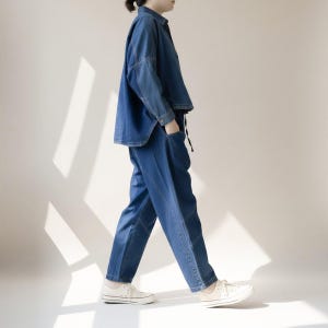 May include: A denim outfit featuring a button-down shirt and matching pants. The shirt has a relaxed fit, and the pants have a unique design. The outfit is a medium blue color. White sneakers complete the look.