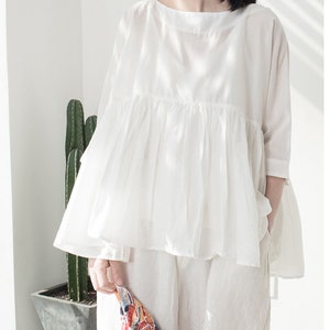 Könnte beinhalten: Eine weiße, lockere Bluse mit geraffter Taille und 3/4-Ärmeln. Die Bluse ist aus einem leichten Stoff gefertigt und hat eine lockere Passform.