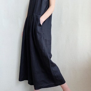 May include: A black linen jumpsuit with thin straps and a wide leg. The jumpsuit has a relaxed fit and is made of a lightweight fabric. The model is wearing the jumpsuit with a white tank top underneath and sandals.