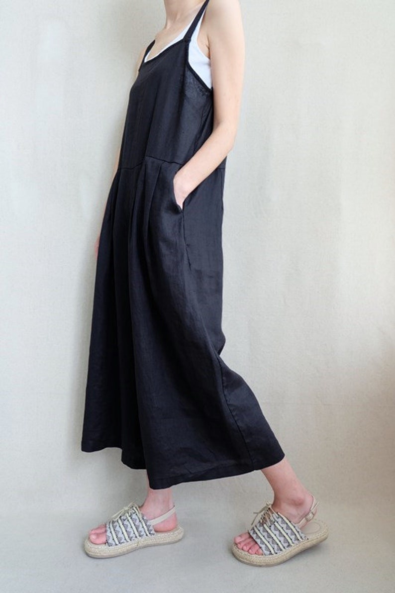 May include: A black linen jumpsuit with thin straps and a wide leg. The jumpsuit has a relaxed fit and is made of a lightweight fabric. The model is wearing the jumpsuit with a white tank top underneath and sandals.